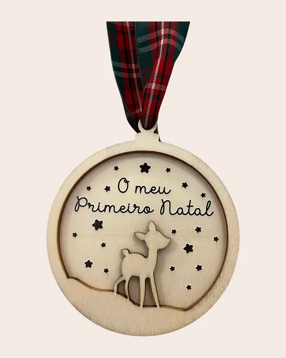 Enfeite redondo em madeira crua, vazado, com a frase «O meu Primeiro Natal» em letra corrida, um veadinho e estrelas, pendurado por fita xadrez verde e vermelha.