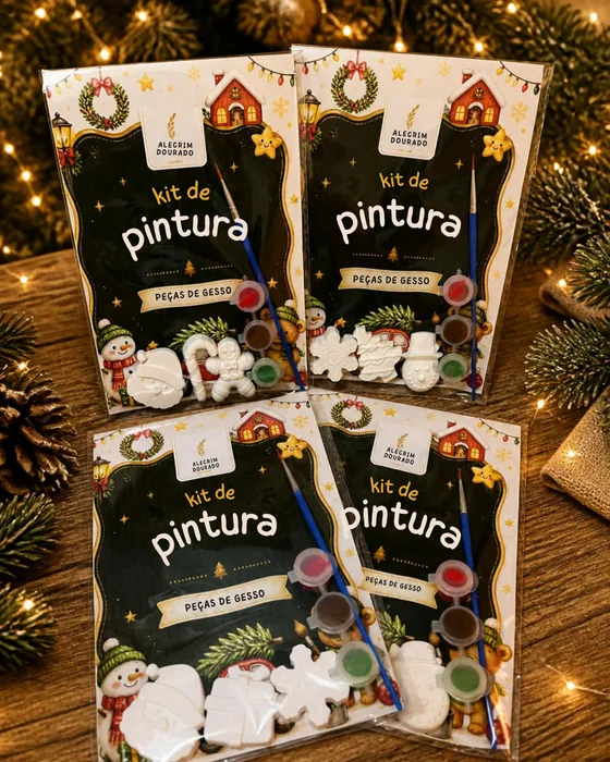 Quatro sacos de kit de pintura da Alecrim Dourado pousados numa mesa de madeira, entre luzes quentes, ramos de pinheiro e uma pinha. Cada saco traz peças de gesso brancas por pintar — Pai Natal, boneco de neve, floco de neve, bengala doce, casinha —, três tintas e um pincel, e diz «kit de pintura · peças de gesso».